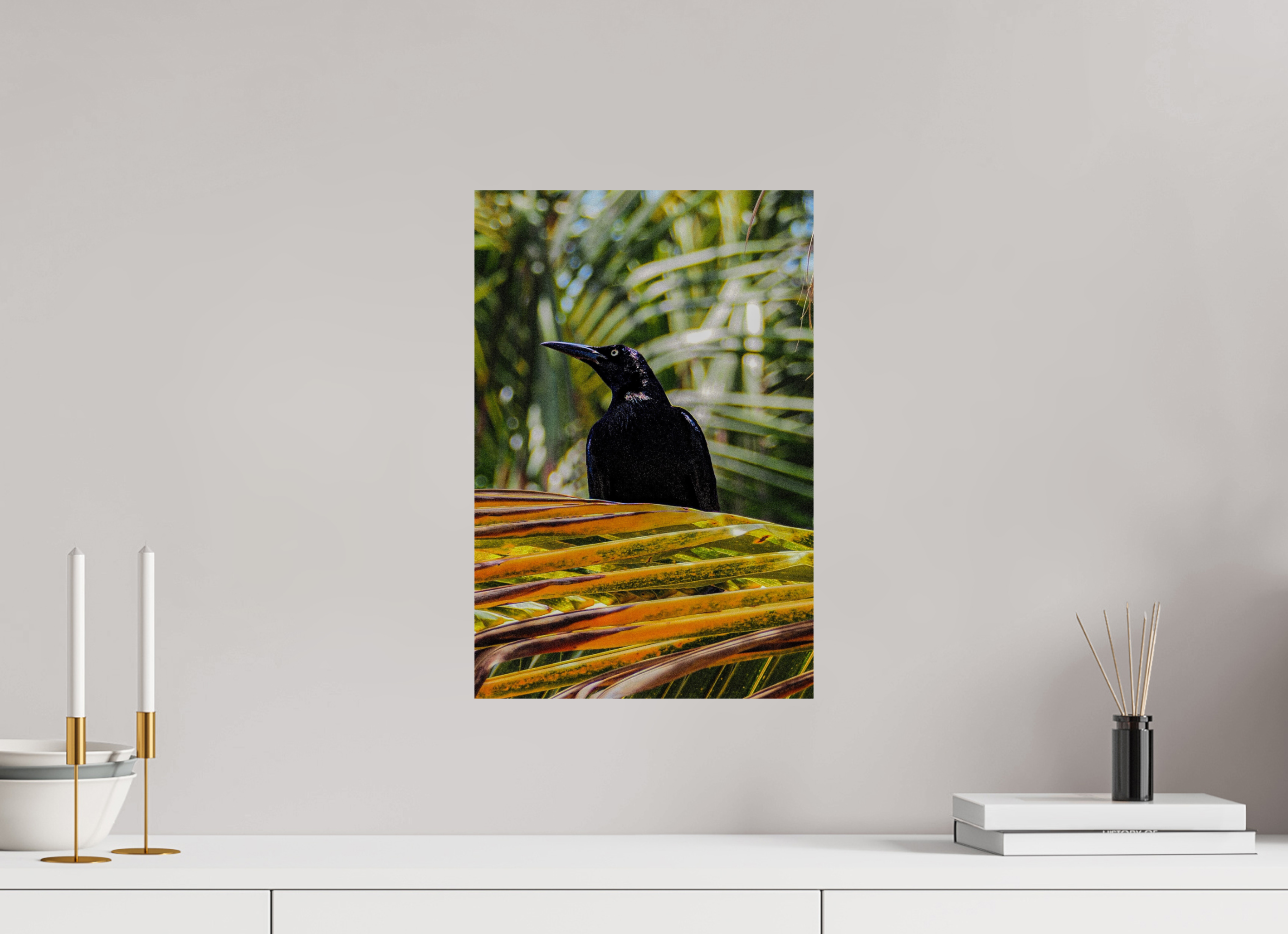 Unframed artwork of a black bird on a branch with green leaves on a white wall.